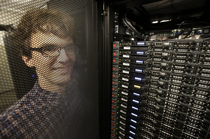 In this Wednesday, June 22, 2016, photo, Massachusetts Institute of Technology researcher Carl Vondrick looks through a protective door while standing next to a computer server cluster, right, on the MIT campus, in Cambridge, Mass. MIT says a computer that binge-watched TV shows such as “The Office,” “Big Bang Theory” and “Desperate Housewives” learned how to predict whether the actors were about to hug, kiss, shake hands or slap high-fives, a breakthrough that eventually could help the next generation of artificial intelligence function less clumsily. (AP Photo/Steven Senne)