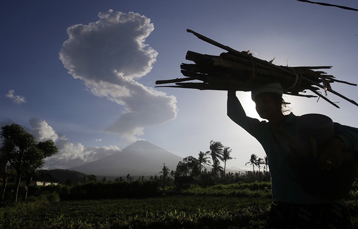 A woman carries wood past as the Mount Agung spews ash and smoke in the background in Karangasem, Bali, Indonesia on July 5, 2018. (AP Photo/Firdia Lisnawati)