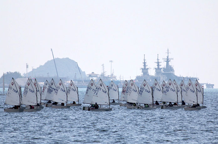 Optimist craft assemble at the start line with Royal Thai Navy warships in the background.