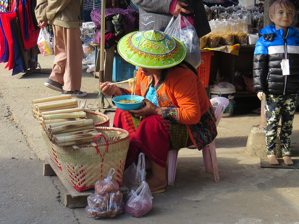 A local vendor enjoys some noodles while waiting for customers. The hats are handmade and embroidered during the rainy season.