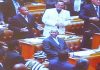 In this photo displayed during his presentation, H.E. Ambassador Geoff Doidge (white suit) stands behind a sitting Nelson Mandela during the time he was in Parliament and Nelson Mandela was President.