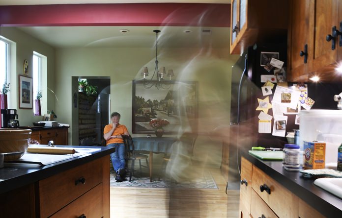 In this Friday, July 13, 2018, photo made with a slow shutter speed, Anne Hunt goes to measure ingredients before adding them to her dough while her husband, Bruce, keeps an eye on her in their home in Chicago. Anne, who once ran a Chicago cooking school, has to separate the ingredients and mixing to two different sections of the kitchen to prevent any errors. She was diagnosed with Alzheimer’s in 2016. (AP Photo/Annie Rice)